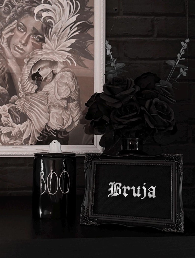May include: A black and white gothic-themed display featuring a framed artwork, a black ceramic jar with "BOO" in white, a framed cross-stitch with the word "Bruja", and a bouquet of black roses. The background includes a black brick wall.