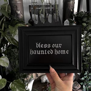 Puede incluir: Un letrero enmarcado en negro con el texto "bless our haunted home" en una fuente gótica. El letrero se sostiene frente a un estante con botellas decorativas, tubos de vidrio con plantas y vegetación. La estética general es oscura y gótica.