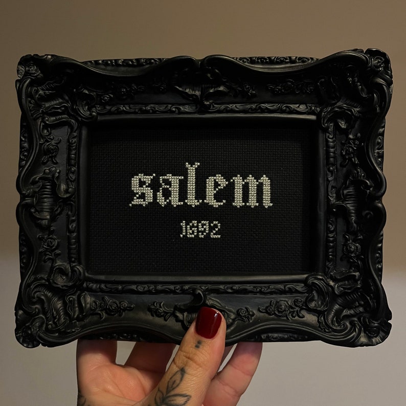 May include: A black framed cross-stitch with white embroidery that reads "Salem 1692".