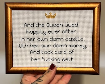 Pode incluir: Um bordado de ponto cruz emoldurado com uma moldura dourada e uma coroa dourada no topo. O bordado diz "And the Queen lived happily ever after. In her own damn castle. With her own damn money. And took care of her fucking self."