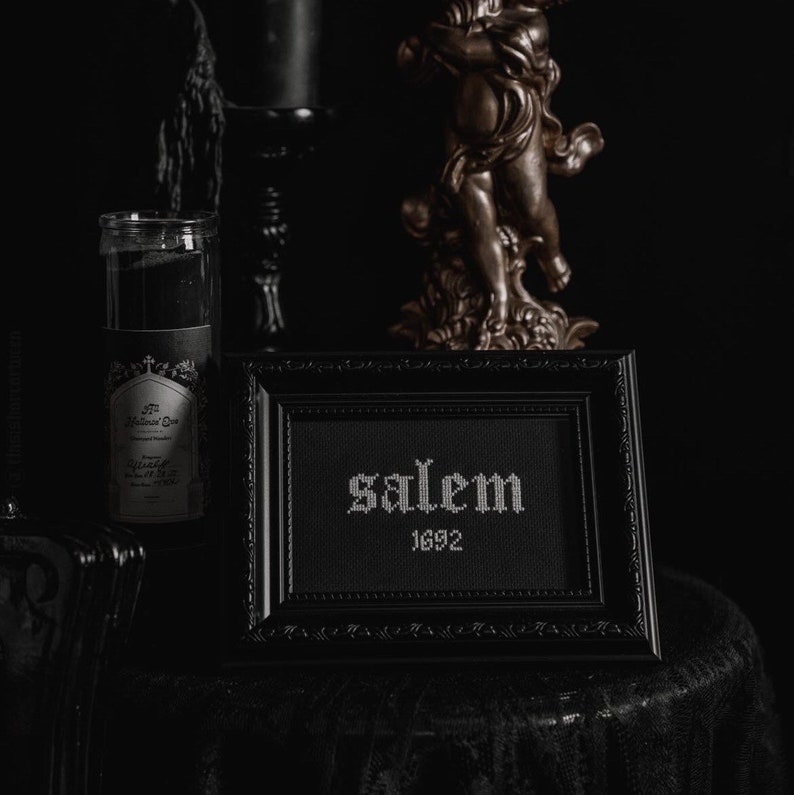 May include: A black framed picture with white embroidery that reads "Salem 1692". The picture is sitting on a black table cloth with a gold cherub statue in the background.
