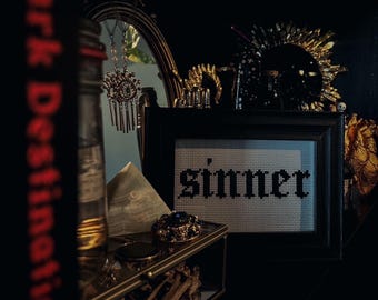 Pode incluir: Uma natureza morta escura e melancólica com um ponto de cruz enquadrado com a palavra "sinner" em letras góticas. A moldura é preta e o ponto de cruz é branco com letras pretas. A moldura está sentada em uma prateleira com outros itens decorativos, incluindo um frasco de vidro, um cristal e uma caixa dourada.
