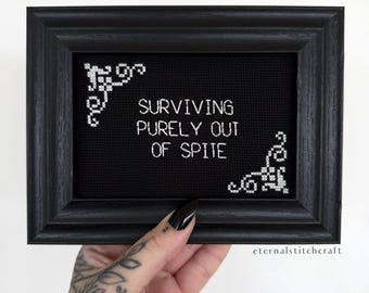 Pode incluir: Arte em ponto cruz com moldura preta com o texto branco "SURVIVING PURELY OUT OF SPITE". O ponto cruz está sobre um fundo preto com detalhes decorativos brancos nos cantos. A moldura é preta escura fosca.