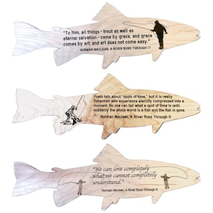 Puede incluir: Tres adornos de madera con forma de pez con un patrón de veta de madera. Cada adorno presenta una ilustración en blanco y negro de una persona pescando y una cita del libro de Norman Maclean "A River Runs Through It".
