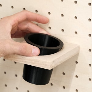 May include: A black plastic cup holder mounted on a light wood pegboard wall.