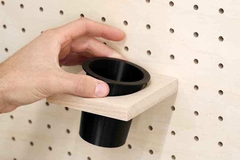 May include: A black plastic cup holder mounted on a light wood pegboard wall.