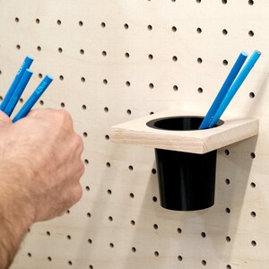 May include: A wooden wall organizer with a black plastic cup attached to it. The organizer is mounted on a pegboard wall. Three blue pencils are being held in a hand.