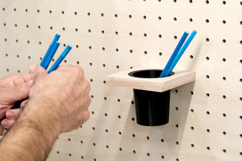 May include: A wooden wall organizer with a black plastic cup attached to it. The organizer is mounted on a pegboard wall. Three blue pencils are being held in a hand.