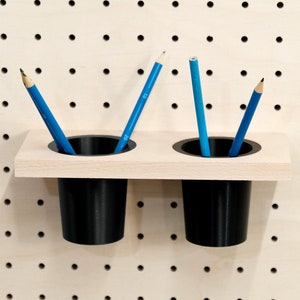 May include: Two black pencil holders mounted on a light wood shelf. The holders are attached to a pegboard wall. There are three blue pencils in the holders.