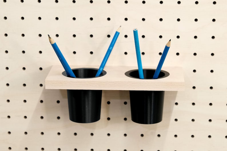 May include: Two black pencil holders mounted on a light wood shelf. The holders are attached to a pegboard wall. There are three blue pencils in the holders.