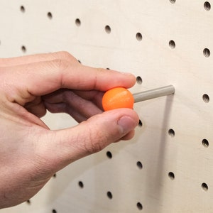 May include: A hand holding an orange ball on a pegboard with a silver peg. The pegboard has a grid of evenly spaced holes.