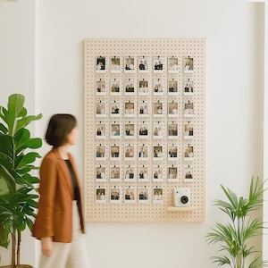 Peut inclure: Un présentoir en bois perforé avec de nombreuses petites photographies attachées. Un petit appareil photo blanc est posé sur une étagère sous les photos. Une personne passe devant l'écran, avec des plantes vertes au premier plan.