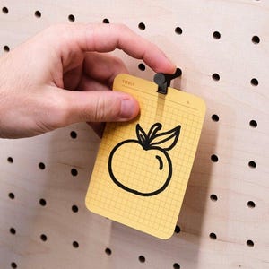 Peut inclure: Une carte rectangulaire jaune avec un motif quadrillé et un dessin au trait noir représentant une pomme au centre. La carte porte le mot TITOLO imprimé en haut et est maintenue par une petite pince en métal noir fixée sur un panneau perforé.