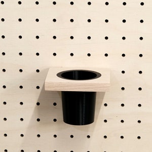 May include: A black plastic cup holder mounted on a wooden pegboard. The holder is attached to a square wooden base.