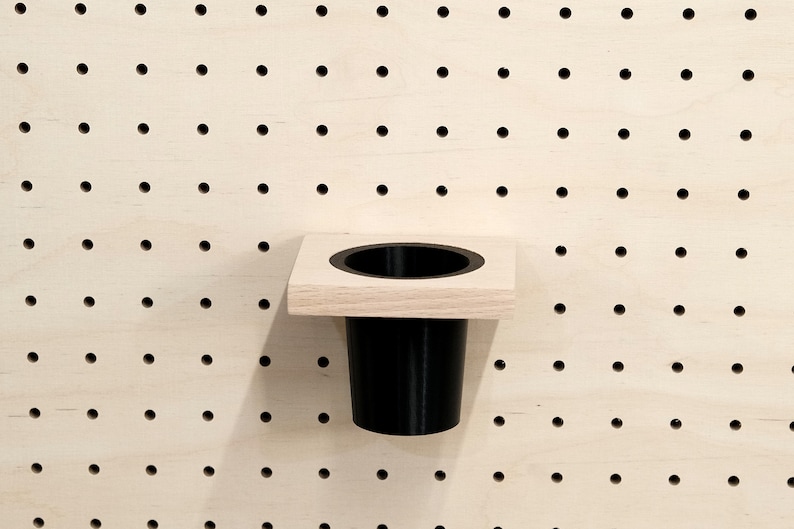 May include: A black plastic cup holder mounted on a wooden pegboard. The holder is attached to a square wooden base.