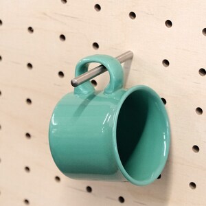 Peut inclure: Une tasse en céramique turquoise avec un crochet métallique accroché à un mur en panneau perforé.