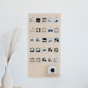 Peut inclure: Un panneau perforé en bois clair avec de nombreuses petites photos attachées. Un appareil photo instantané blanc est posé sur une petite étagère en bas. Des vases décoratifs et une plante sont au premier plan.