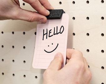 Könnte beinhalten: Eine rosa Notizkarte mit dem Wort "HELLO" in schwarzem Stift geschrieben und einem darunter gezeichneten Smiley. Die Karte ist mit einer schwarzen Klammer an einer Lochwand befestigt.