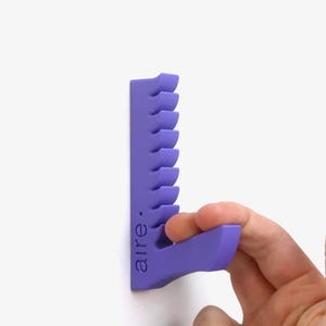 Peut inclure: Un organisateur mural violet pour outils capillaires. L'organisateur comporte une série d'emplacements incurvés pour maintenir les outils et le mot "aire." imprimé sur le côté. Une main tient le bas de l'organisateur.