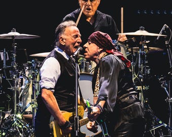 Könnte beinhalten: Zwei Männer auf der Bühne, einer mit einem Mikrofon und einer gelben E-Gitarre, der andere mit einem schwarzen Bandana und einer Gitarre, ein dritter Mann im Hintergrund mit einem Schlagzeugstock.