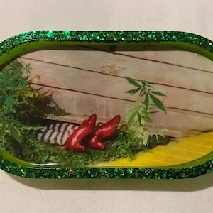 Puede incluir: Una bandeja ovalada de resina con un borde verde brillante. El interior de la bandeja presenta una escena con zapatos rojos, calcetines a rayas y plantas de cannabis. El fondo incluye una valla de madera y un camino de ladrillos amarillos.
