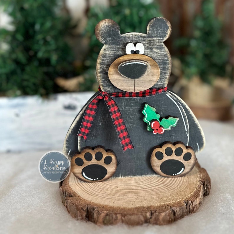 K&ouml;nnte beinhalten: Eine B&auml;renfigur aus Holz mit abgenutztem schwarzem Finish, einem rot-schwarzen Schottenkaro-Schal und einem gr&uuml;nen Stechpalmenblatt-Akzent mit roten Beeren. Der B&auml;r sitzt auf einem Holzscheiben-Sockel und zeigt das Logo L. Krupp Kreations.