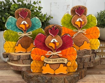 May include: Three wooden turkey figurines standing on natural wood slice bases. Each turkey features a colorful fan-shaped tail with leaf patterns, a red wattle, and orange feet. The center turkey holds a banner that reads Happy Thanksgiving.