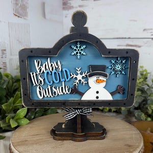 May include: A decorative wooden sign featuring a blue background with the text Baby It's Cold Outside in white and blue lettering. The sign includes a white snowman with a black hat and orange nose, three light blue snowflakes, and a black and white gingham bow.