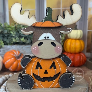 May include: A wooden moose figurine featuring a brown head with antlers, wearing a green-stemmed orange pumpkin as a hat. The moose sits atop a larger orange jack-o'-lantern with black cut-out eyes and a mouth, accented with white dots.
