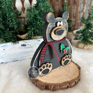 K&ouml;nnte beinhalten: Eine schwarze B&auml;renfigur aus Holz mit abgenutztem Finish, verziert mit einem rot-schwarz karierten Band um den Hals und einem gr&uuml;nen Stechpalmenblatt-Akzent mit roten Beeren. Der B&auml;r steht auf einem runden Sockel aus einer Holzscheibe.