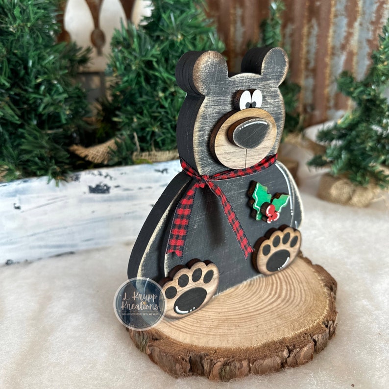 K&ouml;nnte beinhalten: Eine schwarze B&auml;renfigur aus Holz mit abgenutztem Finish, verziert mit einem rot-schwarz karierten Band um den Hals und einem gr&uuml;nen Stechpalmenblatt-Akzent mit roten Beeren. Der B&auml;r steht auf einem runden Sockel aus einer Holzscheibe.