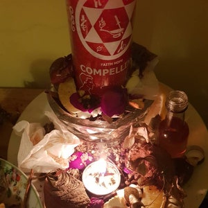 Puede incluir: Una vela roja con una estrella blanca de seis puntas y las palabras "Faith Hope Compelling" en el lateral. La vela está rodeada de flores secas, fruta y una pequeña botella de vidrio.