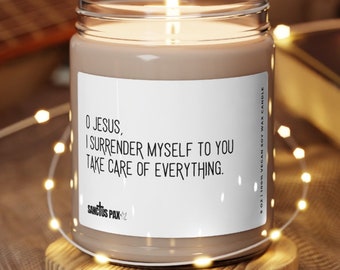 May include: A brown soy wax candle with a white label that reads "O JESUS, I SURRENDER MYSELF TO YOU TAKE CARE OF EVERYTHING." The candle is on a wooden base and surrounded by string lights.