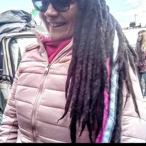 Könnte beinhalten: Eine Frau mit langen, dunklen Dreadlocks mit rosa und weißen Akzenten trägt eine rosafarbene Steppjacke und eine Sonnenbrille. Sie lächelt und schaut nach rechts.