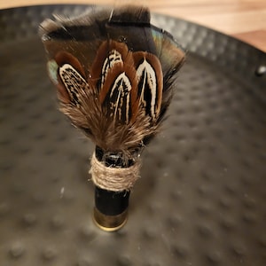 May include: A decorative feather arrangement. The feathers are brown, black, and white, with a touch of iridescent green. The base is a black cylinder with a gold-colored rim, wrapped with twine.
