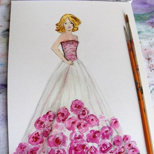 Pode incluir: Pintura em aquarela de uma ilustra&ccedil;&atilde;o de moda. A obra de arte retrata uma mulher em um vestido rosa e branco adornado com rosas rosas. A mulher tem cabelo loiro e est&aacute; em p&eacute; com uma m&atilde;o na cintura. Pinc&eacute;is e uma paleta de aquarela no fundo.