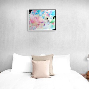 May include: Abstract art print in a black frame. The artwork features a colorful mix of pastel hues, including pink, blue, yellow, and green, with black accents. The painting is displayed above a bed with white bedding and pillows.