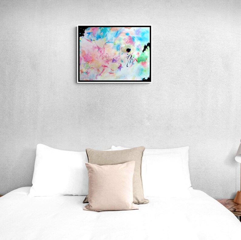 May include: Abstract art print in a black frame. The artwork features a colorful mix of pastel hues, including pink, blue, yellow, and green, with black accents. The painting is displayed above a bed with white bedding and pillows.