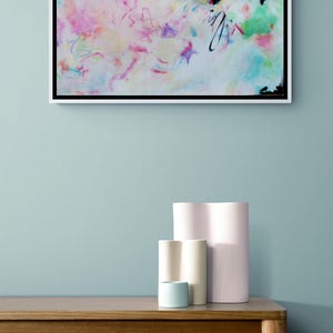 May include: Abstract art piece in a white frame, featuring a vibrant mix of colors including pink, blue, and green. Below, a wooden table displays a stack of books and a collection of decorative vases in various sizes and colors.