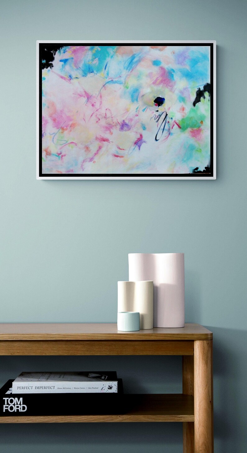 May include: Abstract art piece in a white frame, featuring a vibrant mix of colors including pink, blue, and green. Below, a wooden table displays a stack of books and a collection of decorative vases in various sizes and colors.