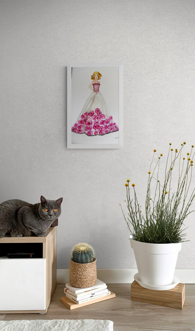 Pode incluir: Uma pintura em aquarela emoldurada de uma mulher em um vestido rosa e branco adornado com rosas cor-de-rosa. Um gato cinza com olhos amarelos est&aacute; sentado em um arm&aacute;rio de madeira. Um cacto em uma cesta e uma planta em vaso tamb&eacute;m s&atilde;o vis&iacute;veis.