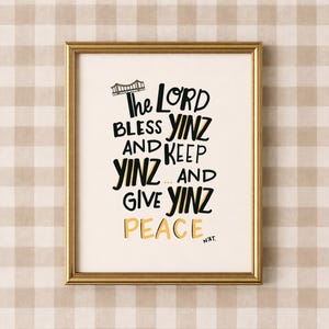 May include: A framed wall art print featuring a gold metallic frame and a cream-coloured background. The text reads: The LORD BLESS YINZ AND KEEP YINZ... AND GIVE YINZ PEACE N'AT. A small black line drawing of a bridge sits above the text.