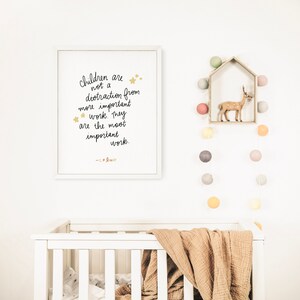 May include: A white framed print with a black handwritten quote that reads "Children are not a distraction from more important work. They are the most important work. -  C.S. Lewis" with gold stars. The print is hanging on a white wall with a white crib and a string of colorful felt balls hanging on the wall.