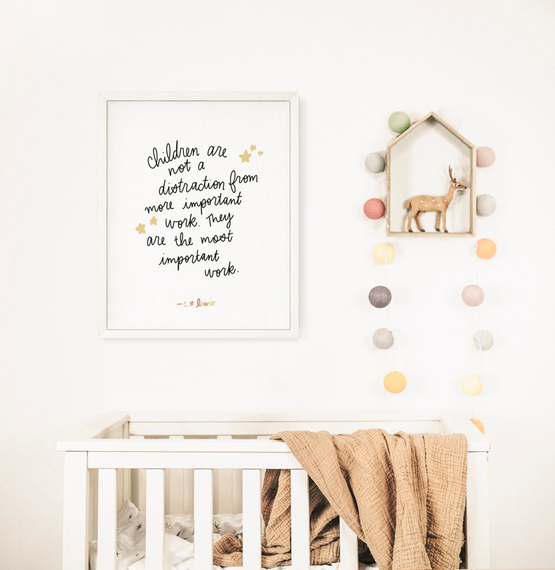 May include: A white framed print with a black handwritten quote that reads "Children are not a distraction from more important work. They are the most important work. -  C.S. Lewis" with gold stars. The print is hanging on a white wall with a white crib and a string of colorful felt balls hanging on the wall.