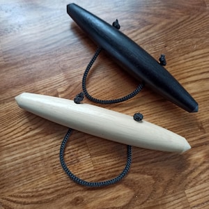 May include: Two wooden yarn swift tools. One is black, the other is natural wood. Each has a rounded, elongated shape and is connected to a black cord. The tools are designed for winding yarn.