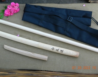 May include: Two wooden practice swords, or bokken, with a black carrying case. The swords are made of light wood and have a natural finish. The case has a shoulder strap and a drawstring closure. The swords are labelled with Japanese characters.