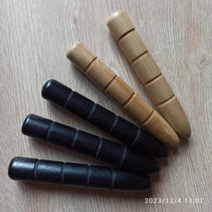 以下が含まれることがあります： 木製のマッサージツールが4つ。2つは薄茶色、2つは黒色です。各ツールは丸みを帯びた端と、一連の彫刻された水平線を持っています。ツールは薄茶色の木製の表面に配置されています。