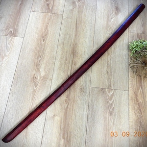 Puede incluir: Una funda de espada de madera larga, delgada y cilíndrica con un acabado teñido de color marrón rojizo oscuro. La superficie muestra un patrón de veta de madera visible a lo largo de toda su longitud, estrechándose hacia el extremo.