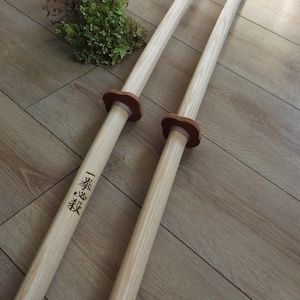 Può includere: Due lunghe spade da allenamento in legno con venature chiare, guardie circolari marroni e un anello rosa chiaro. Una spada presenta i caratteri kanji neri 一拳必殺 incisi sull'impugnatura. Le spade sono lisce e di forma cilindrica.