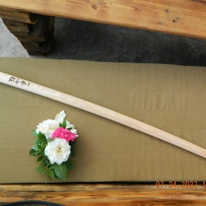 Peut inclure: Un sabre d'entraînement en bois, ou bokken, avec une finition brun clair. Le sabre est posé sur une surface en tissu beige avec un bouquet de roses blanches et roses à gauche. Le sabre porte des caractères japonais inscrits qui se lisent "真剣".